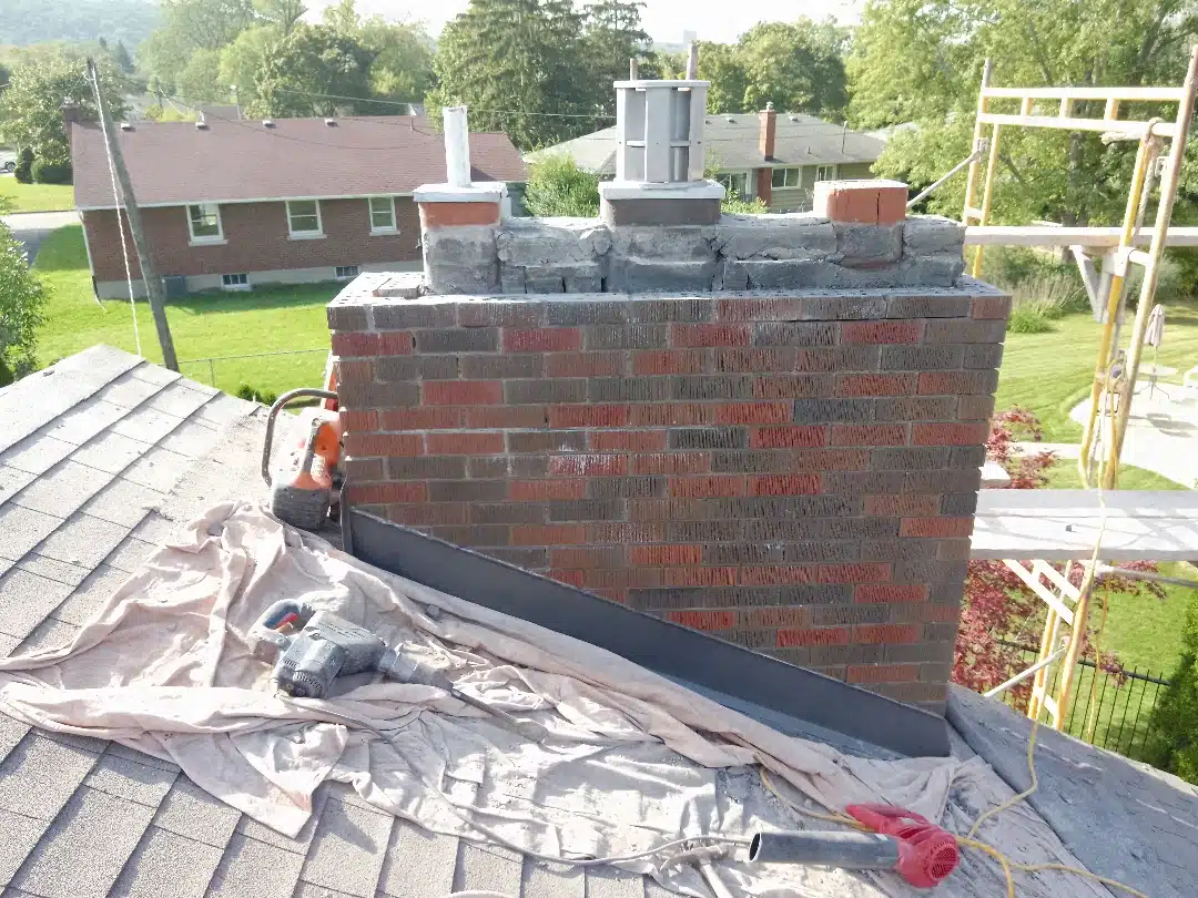 This image shows a partially completed brick chimney on the roof of a residential building The chimney is made of red and grey bricks and has multiple flue pipes protruding from the top There is construction equipment such as a power tool visible on the roof around the chimney The surrounding area includes a grassy lawn and other residential buildings in the background suggesting this is a residential neighborhood setting